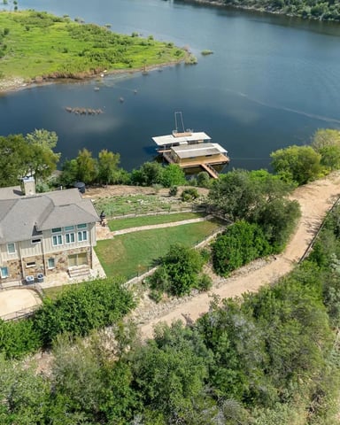 Waterfront house with stone and siding on bluff overlooking river, boat dock, and green lawn, surrounded by trees and rocky shoreline.