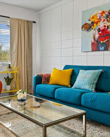 Colorful living room with teal sofa, orange armchair, glass coffee table, and colorful cow artwork on white brick wall