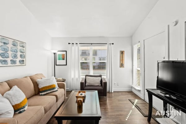 Bright living room with beige sofa, dark armchair, glass coffee table, TV, and windows with white curtains overlooking city buildings