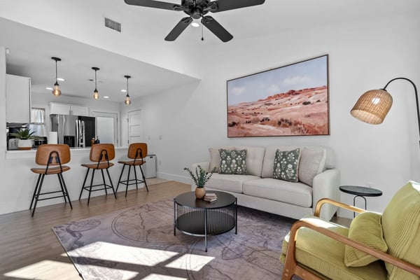 Open-plan living room with beige sofa, green armchair, black round coffee table, and kitchen with brown bar stools in background